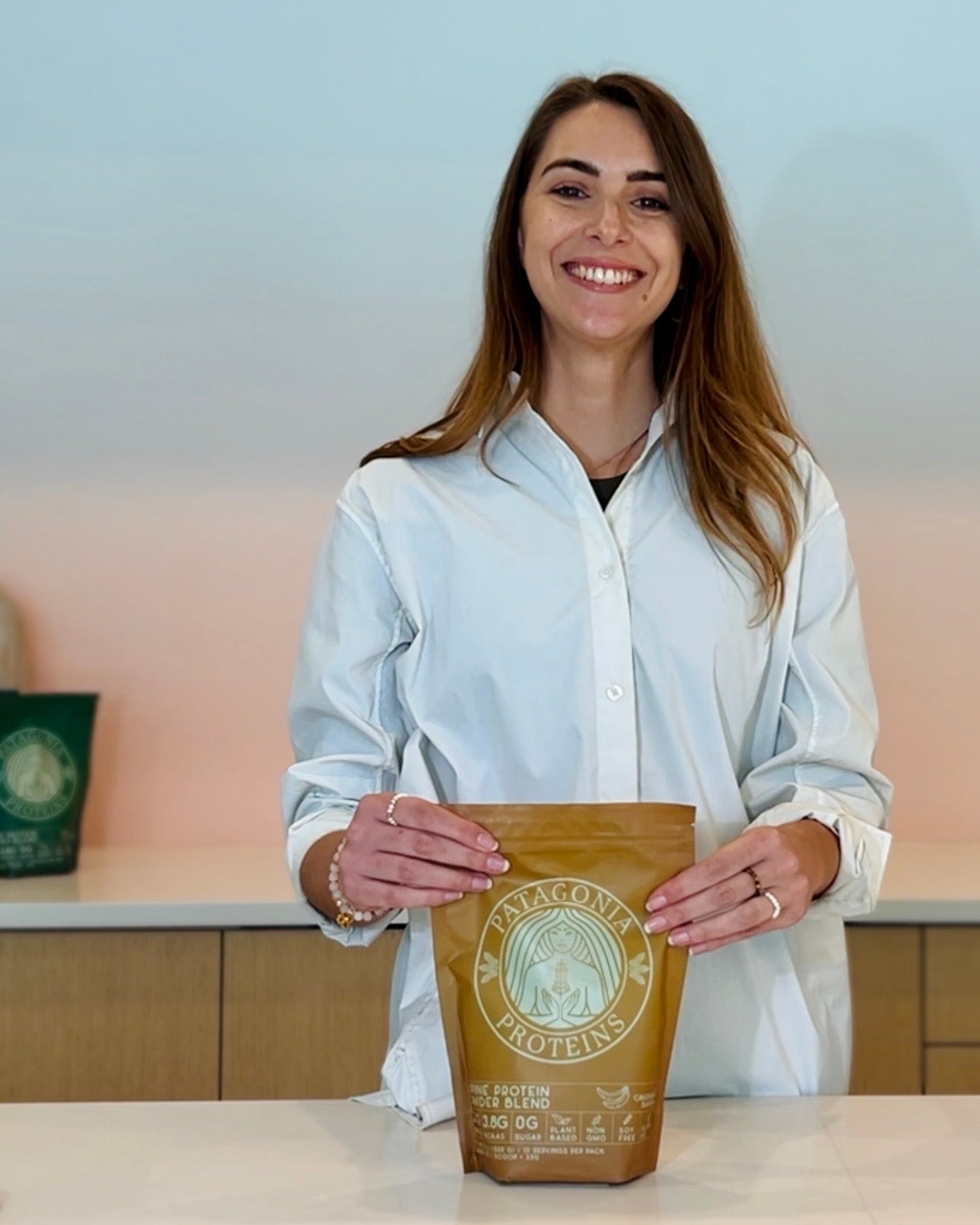 
                  
                    A woman with long brown hair, wearing a white button-down shirt, stands behind a white counter holding a pack of Vegan Cavendish Banana Protein from Patagonia Proteins.
                  
                